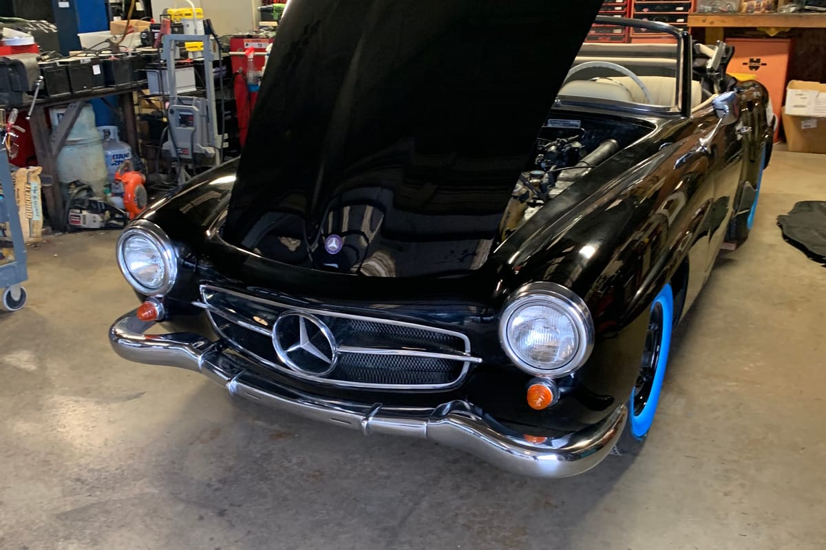 A classic Mercedes-Benz 190SL with its hood raised for repair inside the workshop, with a lift and tools behind